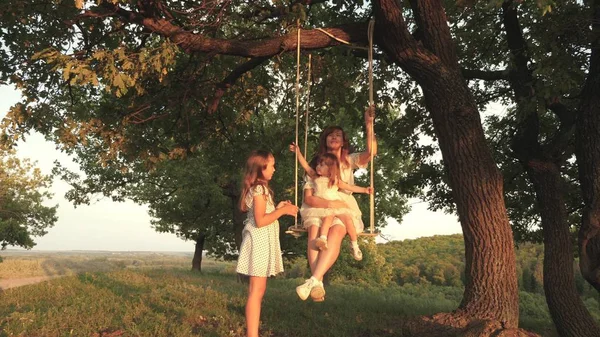 Maman secoue ses filles sur une balançoire sous un arbre au soleil, jouant avec les enfants. mère et bébé se balancent sur une branche de chêne dans la forêt. fille rit, se réjouit. Amusement en famille dans le parc, dans la nature — Photo