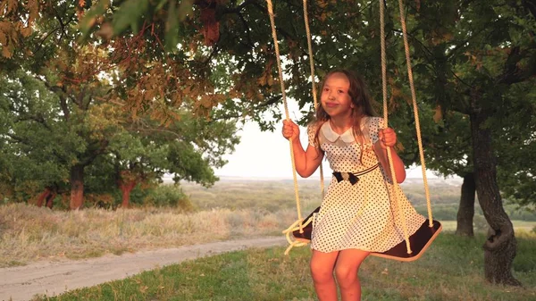 Młoda dziewczyna swinging na huśtawce pod drzewem w słońcu, bawiąc się z dziećmi. Zbliżenie. Rodzinna zabawa w przyrodzie. dziecko jeździ liny huśtawka na gałęzi dębu w parku zachód słońca. śmiech dziewczyna, raduje się. — Zdjęcie stockowe