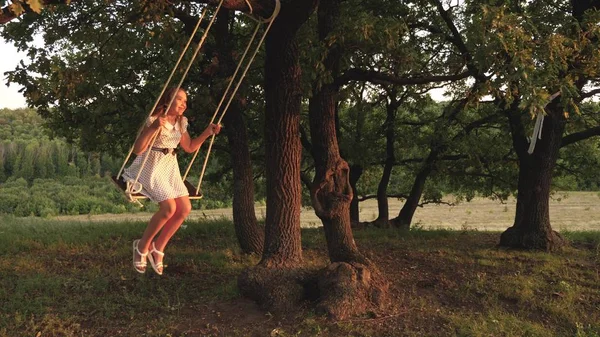 Ung flicka svänger på en gunga under ett träd i solen, leka med barn. Närbild. Familj kul i naturen. barn rider ett rep Swing på en ek gren i parken solnedgången. flickan skrattar, jublar. — Stockfoto