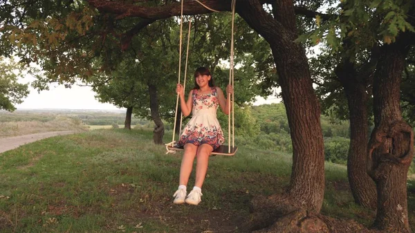 Ung flicka svänga på ett rep Swing på en ek gren. Vacker flicka i en vit klänning i parken. Teen flicka har en flygning på en Swing på en sommarkväll i skogen — Stockfoto