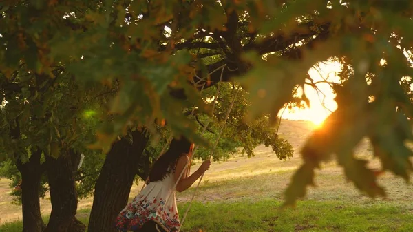 Belle fille balançant sur une balançoire de corde sur une branche de chêne. jeune fille en robe blanche dans le parc. adolescent fille bénéficie d'un vol sur une balançoire un soir d'été dans la forêt . — Photo
