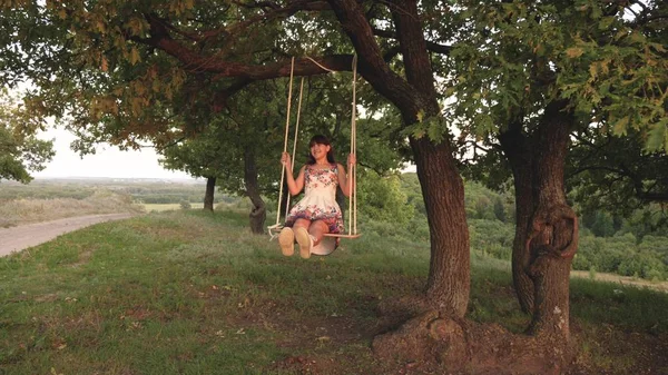 Jong meisje swingende op een touwschommel op een eiken tak. Mooi meisje in een witte jurk in het Park. tiener meisje geniet van een vlucht op een schommel op een zomeravond in het bos — Stockfoto