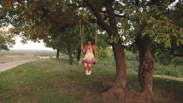 Ung flicka svänga på ett rep Swing på en ek gren. Vacker flicka i en vit klänning i parken. Teen flicka har en flygning på en Swing på en sommarkväll i skogen — Stockfoto
