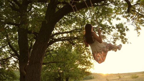 Lycklig tjej svänger på ett rep Swing på en ek gren. ung flicka i en vit klänning i parken. Teen flicka har en flygning på en Swing på en sommarkväll i skogen. — Stockfoto