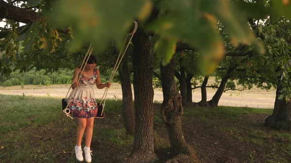 Красивая девочка-подросток наслаждается полетом на качелях летним вечером в лесу. счастливая девушка, качающаяся на веревке на дубовой ветке. Молодая девушка в белом платье в парке . — стоковое фото