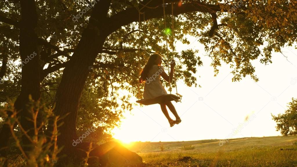child rides a rope swing on an oak branch in forest. girl laughs, rejoices. young girl swinging on a swing under a tree in sun, playing with children. close-up. Family fun in park, in nature.