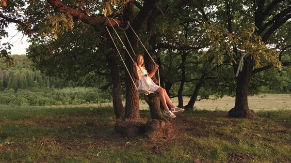 Ung flicka svänger på en gunga under ett träd i solen, leka med barn. Närbild. Familj kul i naturen. barn rider ett rep Swing på en ek gren i parken solnedgången. flickan skrattar, jublar. — Stockfoto