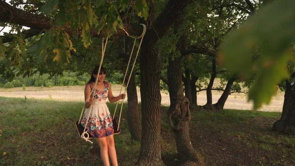 Vacker tonårs flicka njuter av ett flyg på en gunga på en sommarkväll i skogen. lycklig tjej svänger på ett rep Swing på en ek gren. ung flicka i en vit klänning i parken. — Stockfoto