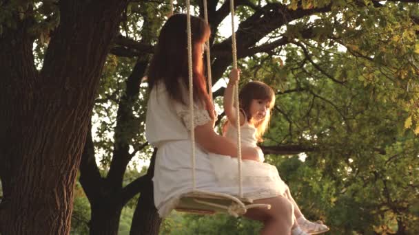 Mutter schüttelt ihre Tochter auf Schaukel unter einem Baum in der Sonne. Nahaufnahme. Mutter und Baby reiten auf einer Seilschaukel auf einem Eichenzweig im Wald. Mädchen lacht, freut sich. Familienspaß im Park, in der Natur. Warmer Sommertag. — Stockvideo
