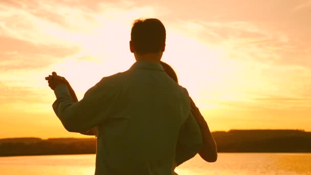 Pareja joven bailando al atardecer en la playa. Hombre y mujer cariñosos bailan en brillantes rayos de sol en el fondo del lago. Feliz chico y chica vals en la noche en el parque de verano . — Vídeos de Stock