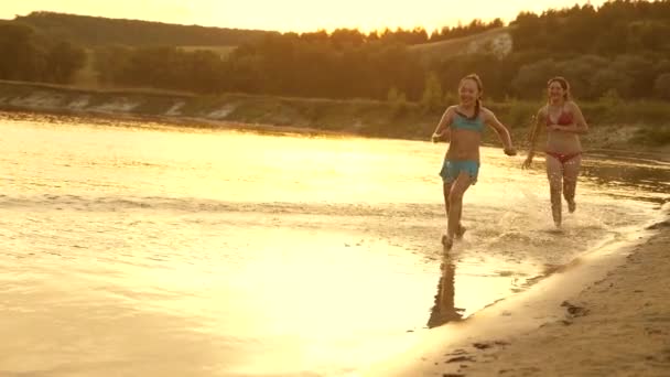 Dziewczyny biegną na piasku wzdłuż brzegu na plaży rozpryskiwania krople wody i śmiech. Szczęśliwych i wolnych nastolatków na wakacjach mają odpocząć o zachodzie słońca. Pracy zespołowej — Wideo stockowe