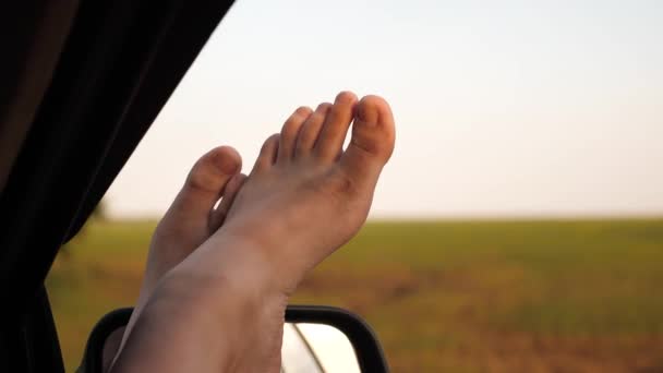 Jonge vrouw houdt van reizen in een auto, het zetten van haar benen uit een open raam. blote voeten van een meisje in een autoruit, schittering van de zon, rijden op een auto op een landweg. vrouw reist met de auto. — Stockvideo