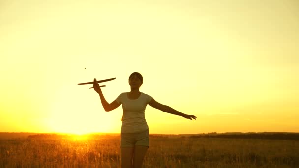Lycklig flicka springer med ett leksaksplan på ett fält i solnedgångens sken. Barn leker leksaksflygplan. Tonåringen drömmer om att flyga och bli pilot. Flickan vill bli pilot och astronaut. Långsamma rörelser — Stockvideo