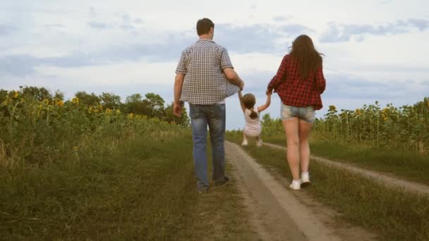 Hija pequeña saltando de la mano de mamá y papá. Familia con niño pequeño camina por la carretera y se ríe al lado del campo de girasoles. Mamá, papá y su hija están descansando juntos fuera de la ciudad en la naturaleza — Vídeo de stock