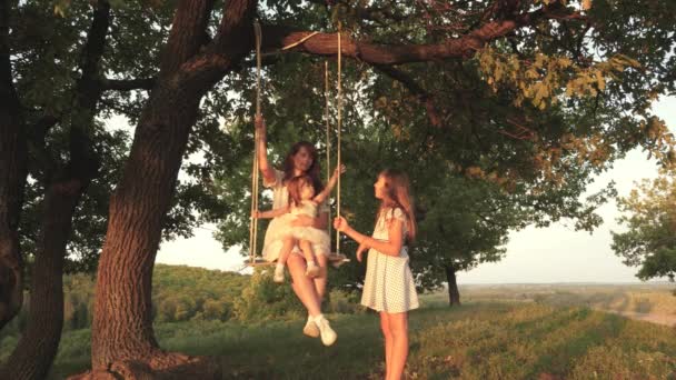 Mother shakes her daughter on swing under a tree in sun. child laughs and rejoices. Mom and baby ride on a rope swing on an oak branch in forest. Family fun in park, in nature. warm summer day. — Stock Video