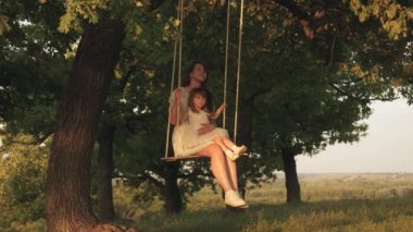 anne ve bebek ormanda bir meşe dalı üzerinde bir ip salıncak binmek. Anne güneşte bir ağacın altında salıncakta kızını sallar. Kız güler, sevinir. Parkta, doğada aile eğlencesi. sıcak yaz günü.