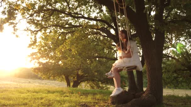 Mamma skakar sin dotter på swing under ett träd i solen. mor och baby rida på ett rep Swing på en ek gren i skogen. Flickan skrattar, jublar. Familj kul i parken, i naturen. varma sommardagen. — Stockvideo