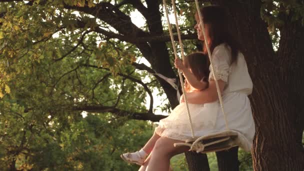 母亲和婴儿骑在森林的橡树树枝上的绳索摆动。女孩笑了，高兴。家庭乐趣在公园，在自然。温暖的夏日。妈妈在阳光下摇摇着女儿在树下。特写. — 图库视频影像
