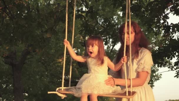 Mother and baby ride on a rope swing on an oak branch in forest. Family fun in park, in nature. Mom shakes her daughter on swing under tree in sun. close-up. child laughs and rejoices. warm summer day — Stock Video