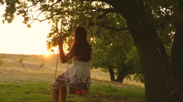 オークの枝にロープを振る若い女の子。公園の白いドレスを着た美少女。十代の女の子は森の夏の夜にブランコでフライトを楽しんでいます — ストック動画