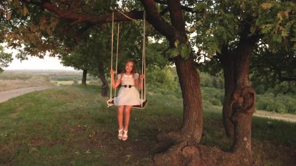 Kind rijdt touwschommel op een eiken tak in het Park de zonsondergang. meisje lacht, verheugt. jong meisje swingende op een schommel onder een boom in de zon, spelen met kinderen. Familie plezier in de natuur. — Stockvideo