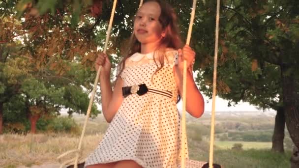 Młoda dziewczyna swinging na huśtawce pod drzewem w słońcu, bawiąc się z dziećmi. Zbliżenie. Rodzinna zabawa w przyrodzie. dziecko jeździ liny huśtawka na gałęzi dębu w parku zachód słońca. śmiech dziewczyna, raduje się. — Wideo stockowe