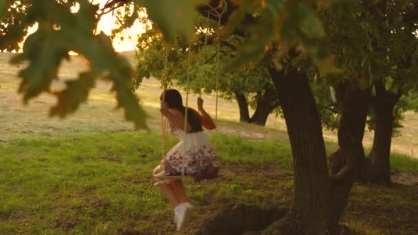 Hermosa chica balanceándose en un columpio de cuerda en una rama de roble. Chica joven con un vestido blanco en el parque. adolescente chica disfruta de un vuelo en un columpio en una noche de verano en el bosque . — Vídeos de Stock