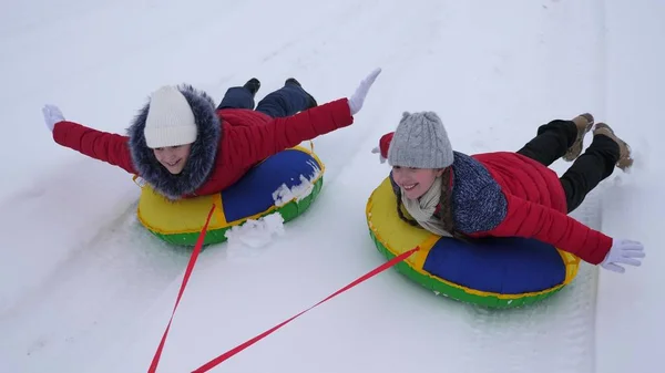 Забавные дети в красных куртках зимой ездят по снегу на санях и надувной снежной трубе и играют в супергероях. Счастливые девушки, отдыхающие в зимнем парке на новогодние праздники. Медленное движение — стоковое фото