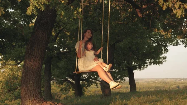 Moeder en baby rijden op een touwschommel op een eiken tak in bos. Moeder schudt haar dochter op schommel onder een boom in de zon. Meisje lacht, verheugt. Familie plezier in Park, in de natuur. warme zomerdag. — Stockfoto