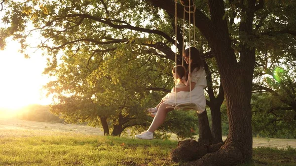 Mamá sacude a su hija en columpio bajo un árbol bajo el sol. madre y el bebé montar en un columpio cuerda en una rama de roble en el bosque. La chica se ríe, se alegra. Diversión familiar en el parque, en la naturaleza. cálido día de verano . — Foto de Stock