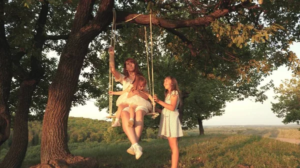 Mamma skakar sin dotter på swing under ett träd i solen. barn skrattar och jublar. Mamma och baby rida på ett rep Swing på en ek gren i skogen. Familj kul i parken, i naturen. varma sommardagen. — Stockfoto