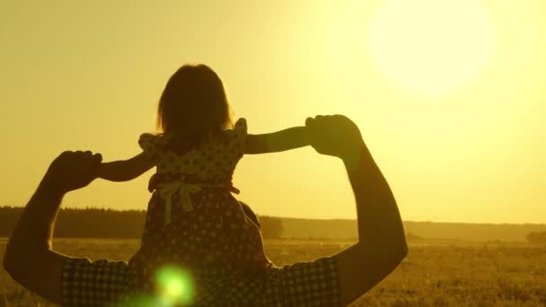 Papá lleva al amado bebé sobre sus hombros caminando alrededor del campo al atardecer. hija pequeña montando con papá sobre sus hombros en el parque. Un niño con sus padres camina por la noche al atardecer del sol. Movimiento lento . — Vídeo de stock