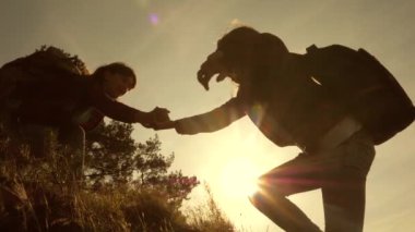 Gezgin kızlar dağa tırmanıyor, birbirlerine yardım eli uzatıyor, tepeye tırmanmaya yardım ediyor. Gün batımında seyahat eden turist ailesi. sırt çantaları ile kız kardeşler güneşte dağa tırmanma seyahat. turistik ekip çalışması