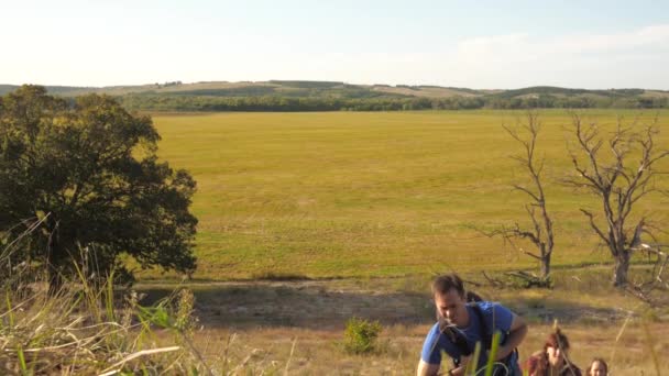 Rodina turistů s dětmi šplhá do kopce. otec má ruku, která pomáhá dětem šplhat po horách. Tati, děti a máma s batohy šplhají po horách na slunci. Turistická spolupráce — Stock video