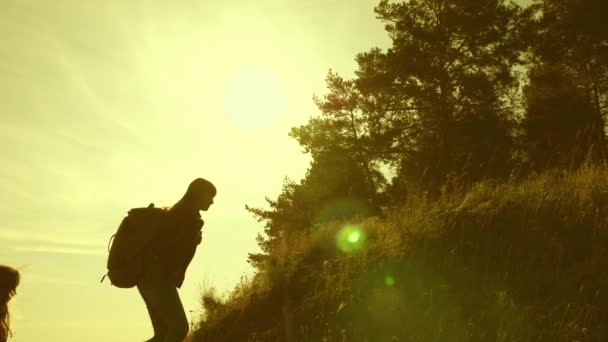 Traveler girls climb mountain, offer each other a helping hand, helping to climb hill. Family of tourists traveling at sunset. sisters with backpacks travel climbing mountain in sun. tourist teamwork — Stock Video