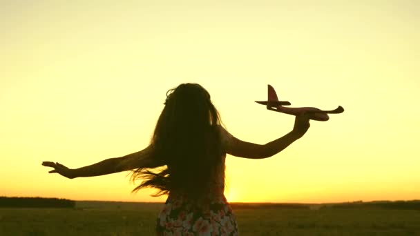 幸せな女の子は日没の光の中でフィールド上のおもちゃの飛行機で実行されます。子供はおもちゃの飛行機で遊ぶ。10代の頃はパイロットになることを夢見ていました。少女はパイロットと宇宙飛行士になりたがっている。スローモーション — ストック動画