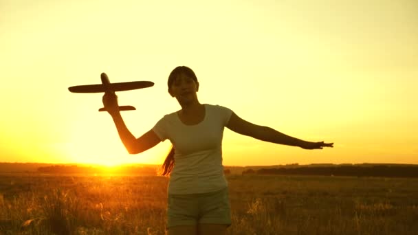 Gelukkig meisje rent met een speelgoedvliegtuigje op een veld bij zonsondergang. kinderen spelen speelgoed vliegtuig. tiener droomt van vliegen en piloot worden. Het meisje wil piloot en astronaut worden. Langzame beweging — Stockvideo