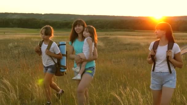 Chicas felices viajan a través del campo en la luz del atardecer. Hiker Girl. familia camina en el prado. niñas viajan con mochilas en una carretera de campo. Una familia feliz de vacaciones. concepto de turismo deportivo — Vídeos de Stock