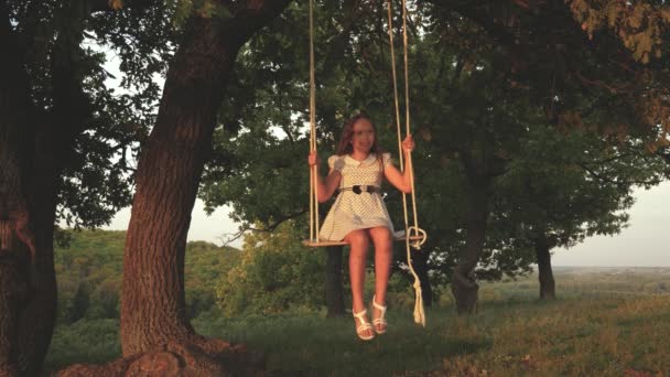 Mladá dívka houpá se na houpačce pod stromem na slunci a hraje si s dětmi. dítě jede po laně na dubové větvi v lese. dívka se směje, radní. Rodinná zábava v parku, v přírodě. — Stock video
