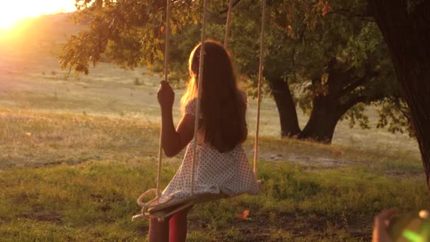 Девочка качается на качелях под солнцем, играет с детьми. крупным планом. Семейное веселье в природе. ребенок ездит на веревке качели на дубовой ветке в парке закат. Девушка смеется, радуется . — стоковое видео