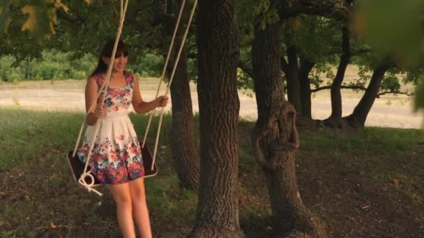 オークの枝にロープスイングで揺れる幸せな女の子。公園の白いドレスを着た若い女の子。十代の女の子は森の夏の夜にブランコでフライトを楽しんでいます. — ストック動画
