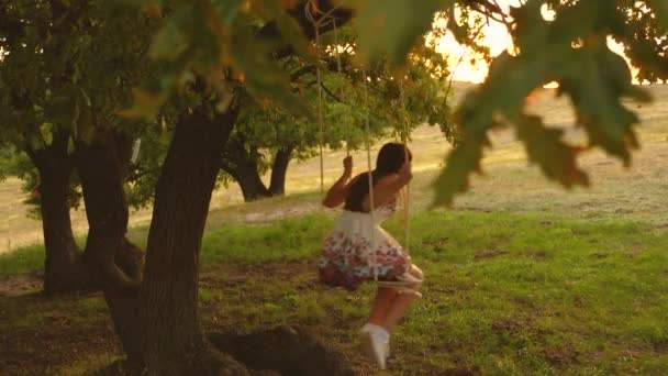 Tiener meisje geniet van een vlucht op een schommel op een zomeravond in het bos. jong meisje swingende op een touwschommel op een eiken tak. Mooi meisje in een witte jurk in het Park. — Stockvideo