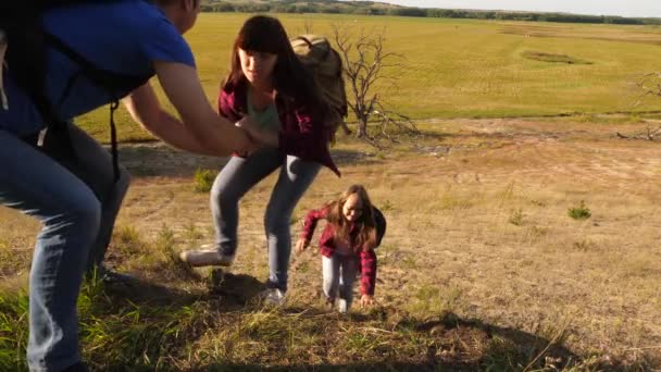 Fadern håller ut sin hand hjälpa barn klättra berg. Familj av turister med barn som reser i solnedgången. Pappa, barn och mamma med ryggsäckar resa klättra berg i solen. turist samarbete — Stockvideo