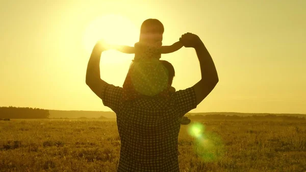 Papá lleva al amado bebé sobre sus hombros caminando alrededor del campo al atardecer. hija pequeña montando con papá sobre sus hombros en el parque. Un niño con sus padres camina por la noche al atardecer del sol. Movimiento lento . — Foto de Stock