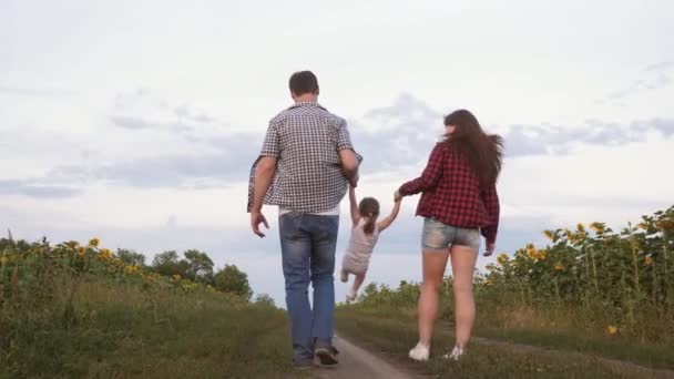 Bambino vola nelle mani di padre e madre. Figlia che salta tenendosi per mano mamma e papa '. Famiglia con bambino piccolo cammina lungo la strada e ride accanto al campo di girasoli . — Video Stock