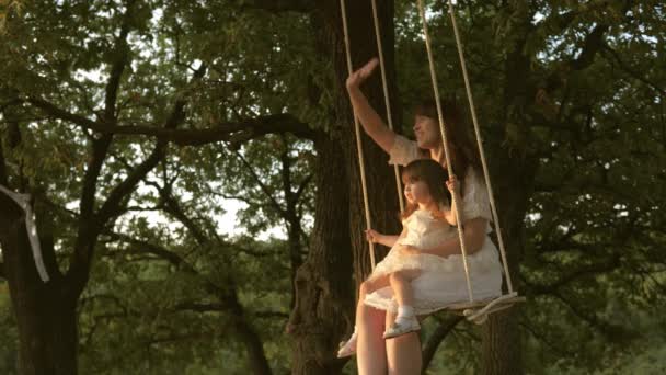Máma otřásá dcerou pod stromem na slunci. Close-up. matka a dítě jedou po laně na dubové větvi v lese. Dívka se směje, radní. Rodinná zábava v parku, v přírodě. teplý letní den.