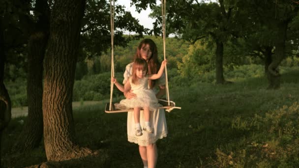 Mamma skakar sin dotter på swing under ett träd i solen. Närbild. mor och baby rida på ett rep Swing på en ek gren i skogen. Flickan skrattar, jublar. Familj kul i parken, i naturen. varma sommardagen. — Stockvideo
