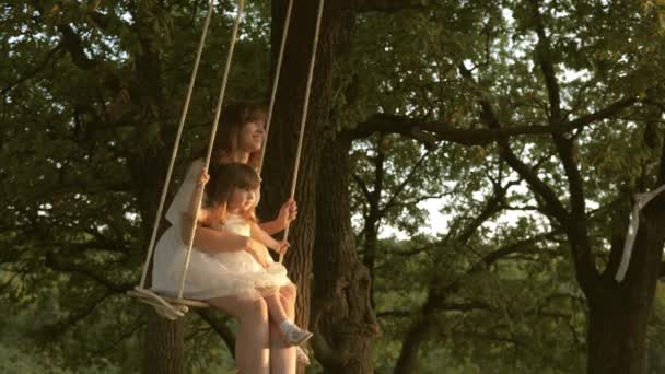 Мать и ребенок едут на веревке качели на дубовой ветке в лесу. Девушка смеется, радуется. Семейное веселье в парке, на природе. теплый летний день. Мама трясет своей дочерью на качелях под деревом на солнце . — стоковое видео