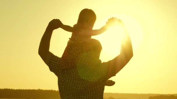 Papá lleva al amado bebé sobre sus hombros caminando alrededor del campo al atardecer. hija pequeña montando con papá sobre sus hombros en el parque. Un niño con sus padres camina por la noche al atardecer del sol. Movimiento lento . — Vídeos de Stock