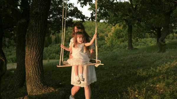 Mamma skakar sin dotter på swing under ett träd i solen. Närbild. mor och baby rida på ett rep Swing på en ek gren i skogen. Flickan skrattar, jublar. Familj kul i parken, i naturen. varma sommardagen. — Stockfoto